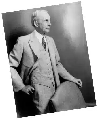 A vintage black-and-white studio portrait of an older Henry Ford dressed in a light-colored three-piece suit, standing and looking off-camera while resting his hand on a leather chair, representing his lasting legacy as a major 20th-century industrialist.