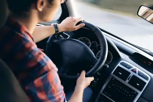 Over-the-shoulder view of a man driving a car with both hands on the black steering wheel, representing vehicle handling and experiencing sudden engine or PCM performance issues while on the road.