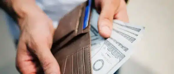 Close-up of a person holding a brown leather wallet and counting one-hundred dollar bills, representing an ECM core deposit refund and cost savings.