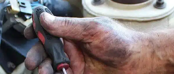 A mechanic's grease-stained hand holding a screwdriver under the hood of a car, representing DIY engine computer troubleshooting and visual inspection.